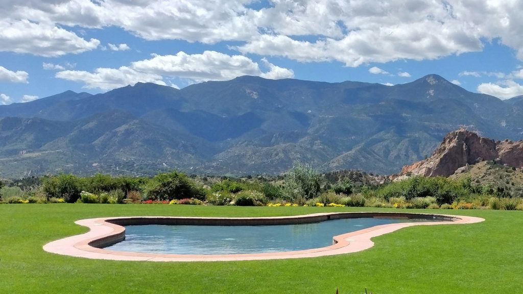 A well-maintained pool surrounded by lush green grass and colorful landscaping, with mountains in the background under a partly cloudy sky, illustrating the challenge of protecting grass from pool water effects.