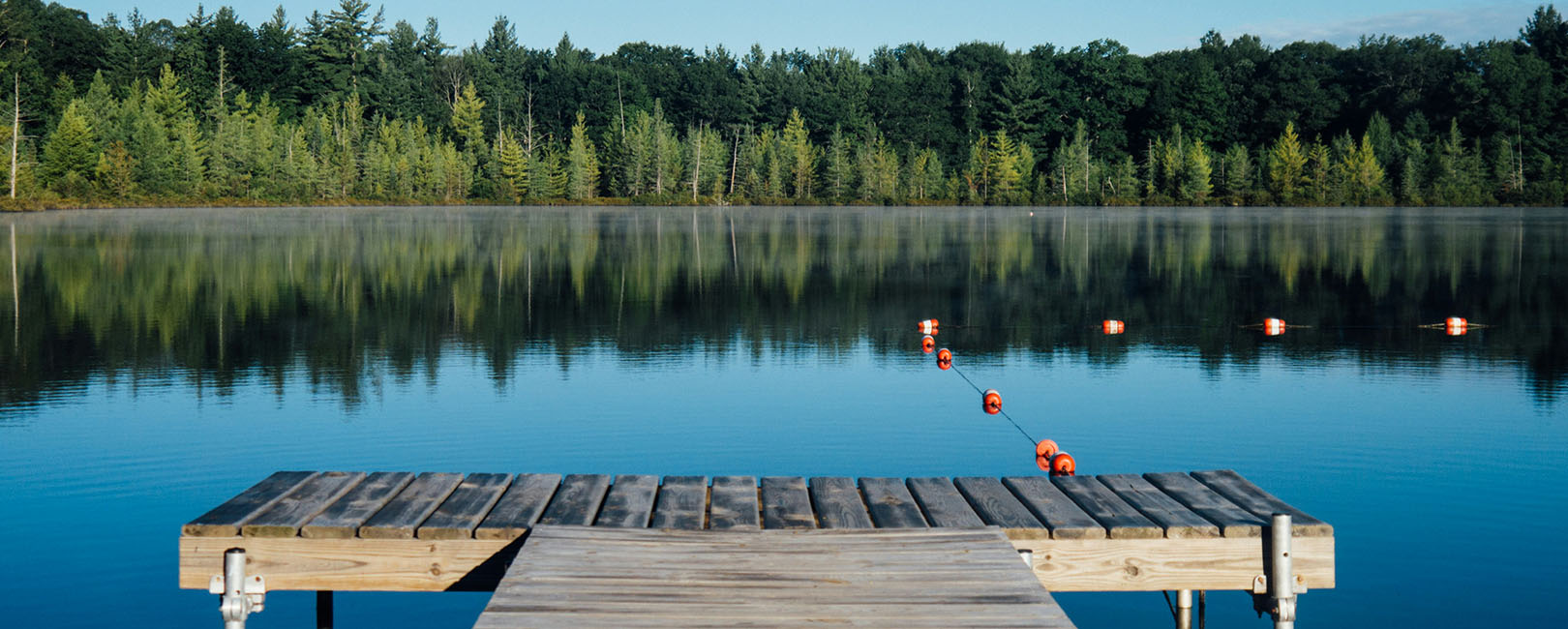 A serene lake with a wooden dock extending into calm, clear water, surrounded by a dense forest under a bright sky, with orange buoys marking a safe swimming area, highlighting the natural beauty and potential safety considerations of lake swimming.